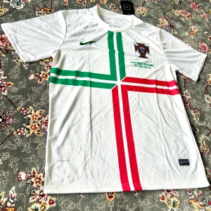 Portugal fotbollströja Nike - Vit Portugal fotbollströja från Nike med grönt och rött korsmönster på framsidan. Broderad Nike-logga i grönt och Portugals landslagsemblem på bröstet. Texten 'Portugal/Espanha 27/06/2012' tryckt under emblemet. Tillverkad i lätt och ventilerande material.