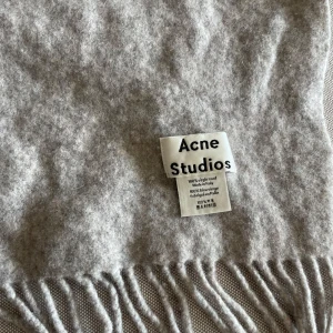 Ljusgrå halsduk från Acne Studios - Säljer en stilren ljusgrå halsduk från Acne Studios i 100% ull. Halsduken har klassiska fransar i kanterna och mäter ca 200x70 cm. Perfekt accessoar för att hålla värmen och lyfta din outfit.