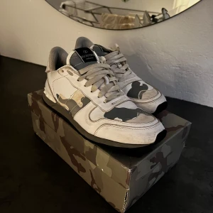 Valentino sneakers med camouflage-mönster - Säljer ett par snygga Valentino sneakers i vitt och grått med coolt camouflage-mönster. Skorna har snörning, rund tå och platt sula. Perfekta för dig som vill sticka ut med exklusiva och trendiga sneakers.
