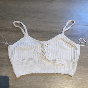 Vit stickad croppad linnetopp från FB Sister - Säljer en vit stickad croppad linnetopp från FB Sister. Toppen har smala axelband och snörning framtill som detalj. Perfekt för varma dagar och enkel att matcha med jeans eller kjol.
