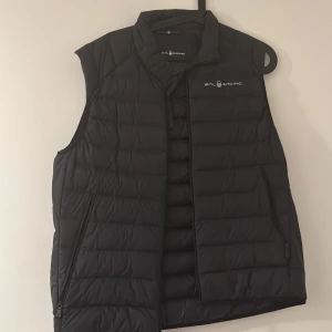 Svart dunväst från Sail Racing - Säljer en svart dunväst från Sail Racing med hög krage och dragkedja framtill. Västen har diskret logga på bröstet och är quiltad för extra värme. Perfekt för lager-på-lager under kyliga dagar. Medium i barn storlek så skulle säga 168-175Cm