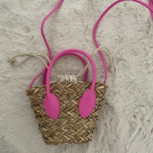 Mini strandväska med rosa detaljer - Superfin mini stråväska med starkt rosa handtag och axelrem. Väskan har en vit tyginsida med dragsko som stängning. Perfekt accessoar för att liva upp din outfit!