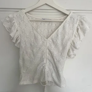 Vit spetsblus med volangärm från Zara - Säljer en söt vit blus från Zara med broderad spets och volangärmar. Blusen har v-ringning, knyt i midjan och ett blommigt hålmönster. Perfekt för vår och sommar!