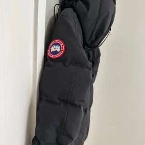 Svart dunjacka från Canada Goose - Säljer en svart dunjacka från Canada Goose med huva och klassisk logotyp på ärmen. Jackan har dragkedja framtill och är perfekt för kalla dagar. Snygg och tidlös modell som håller värmen bra.