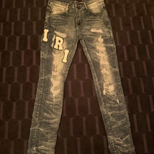 Blå jeans från Amiri - Säljer ett par blå jeans från Amiri med slitningar och stora vita bokstavspatchar på benen. Jeansen har dragkedjegylf och smal passform. Snygga detaljer och cool streetstil. Storlek 30  31