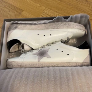 Vita sneakers från Golden Goose med grå stjärna - Säljer ett par vita sneakers från Golden Goose med grå mockadetaljer och en ikonisk grå stjärna på sidan. Skorna har grå snörning och svart hälparti. Perfekta för en trendig och avslappnad stil.