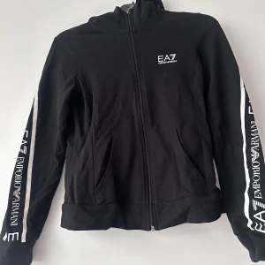 Svart zipjacka från EA7 Emporio Armani - Svart zipjacka med vita detaljer och logga från EA7 Emporio Armani på bröstet och längs ärmarna. Jackan har hög krage, dragkedja framtill och ribbade muddar. Perfekt för en sportig och stilren look. Och mjukisbyxor