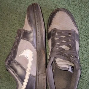 Nike Dunk Low grå/vita sneakers - Säljer ett par Nike Dunk Low sneakers i grått och vitt med klassisk swoosh på sidan. Skorna har snörning, platt sula och rund tå. Perfekta för dig som gillar stilrena och sportiga sneakers.