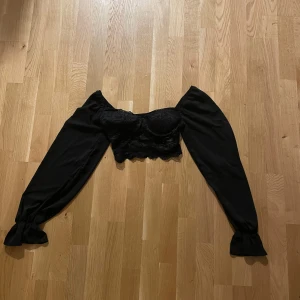 Svart offshoulder topp med spetsdetaljer - Säljer en svart offshoulder topp med långa puffärmar och vacker spets över bysten och nederkanten. Toppen har croppad passform och romantisk känsla, perfekt för att lyfta din outfit.