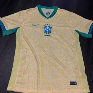 Gul Brasil fotbollströja från Nike - Säljer en gul Brasilien fotbollströja från Nike med blå och gröna detaljer. Tröjan har korta ärmar, V-ringad krage och ett subtilt mönster över hela tyget. Perfekt för fotbollsfans som vill visa sitt stöd för landslaget.