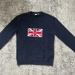 Mörkblå stickad tröja från Pepe Jeans - Mörkblå stickad tröja från Pepe Jeans med ett stort Union Jack-flagga broderat framtill och logga i rött. Klassisk rund halsringning och ribbade muddar vid ärmslut och nederkant. Perfekt för dig som gillar stilrena detaljer.