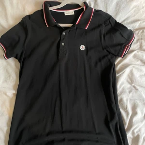 Svart pikétröja från Moncler - Säljer en svart pikétröja från Moncler med klassisk krage och knäppning framtill. Tröjan har röda och vita ränder på krage och ärmslut samt Moncler-logga på bröstet. Perfekt för en stilren och sportig look.