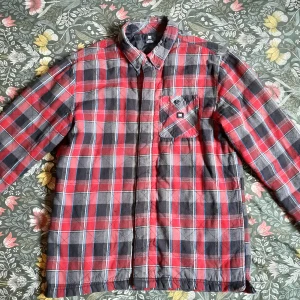 Rutig vadderad skjorta från DC Shoes - Säljer en röd, svart och grå rutig overshirt från DC Shoes. Skjortan har långa ärmar, bröstficka med knapp och quiltat foder på insidan för extra värme. Perfekt att slänga på sig när det är kyligt ute.