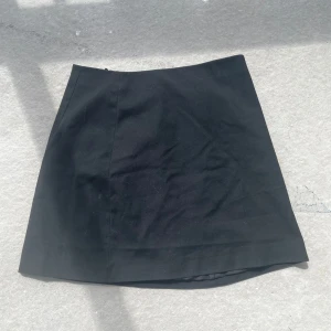 Svart kortkjol från H&M - En stilren svart kortkjol från H&M med dragkedja och knapp bak. Kjolen har en klassisk A-linjeform och är helfodrad för extra komfort. Perfekt att matcha med en snygg topp eller stickad tröja.