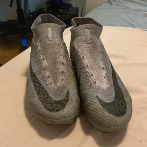 Nike superfly - Säljer ett par gråa Nike fotbollsskor med högt skaft och stora svarta Nike-loggor på sidorna. Skorna har en strumpliknande passform och är designade för att ge bra grepp på planen. Perfekta för dig som vill sticka ut på matchen.