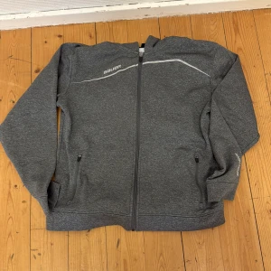 Grå ziptröja från Bauer - Mörkgrå ziptröja från Bauer med hel dragkedja och två fickor framtill. Tröjan har en diskret vit linje över bröstet och Bauer-logga på vänster sida. Perfekt för träning eller vardag.