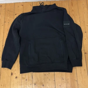 Svart hoodie från Nicce - Säljer en svart hoodie från Nicce med dragsko i huvan och en diskret logotyp på ärmen. Tröjan har en klassisk passform och en stor ficka framtill. Perfekt för en avslappnad stil.
