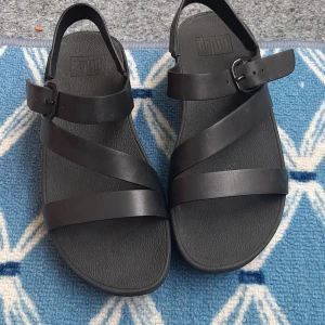 Svarta sandaler från FitFlop - Säljer ett par svarta sandaler från FitFlop med tre remmar och spänne vid ankeln. Skorna har en platt sula och är tillverkade i syntetmaterial. Perfekta för varma dagar och enkel att matcha med olika outfits. Skor används bara några gånger i mycket bara skick. 