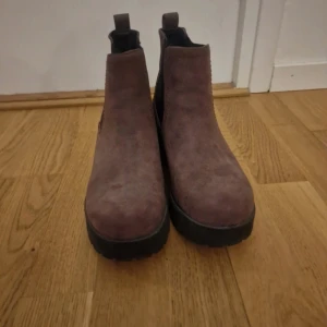 Vinröda chelsea boots med klack - Säljer ett par vinröda chelsea boots i mocka med grov sula och klack. Skorna har elastiska svarta paneler på sidorna och rund tå. Perfekta för dig som vill ha en stilren men ändå edgy look.