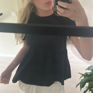 Svart volangtopp från 157 - Säljer en svart topp från 157 med volang nedtill. Toppen har rund halsringning och korta ärmar. Perfekt att matcha med både jeans och kjol för en enkel och stilren look.