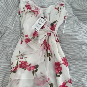 Vit blommig från Zara - Superfin vit omlottklänning från Zara med rosa blommor och spetsdetaljer vid v-ringningen. PRISLAPP KVAR! Klänningen har smala axelband och knyt i midjan för en feminin siluett. Perfekt för sommarens alla tillfällen!
