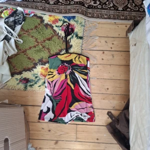 Färgglad axelbandslös topp med blommigt mönster - Säljer en axelbandslös klänning med stort, färgglatt blommönster i rött, rosa, grönt, svart och vitt. Klänningen har en kort längd och är perfekt för dig som gillar att sticka ut med starka färger och mönster.