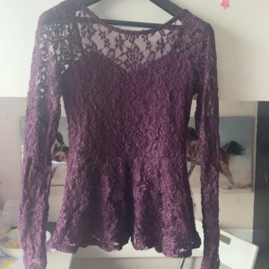 Lila spetsblus med lång ärm - Säljer en lila blus i spets med lång ärm och v-ringning. Blusen har ett vackert blommigt spetsmönster och är figursydd upptill med lätt utställd nederdel. Perfekt för dig som vill ha en romantisk och elegant look. Lite små trådar lite här å där på tröjan 