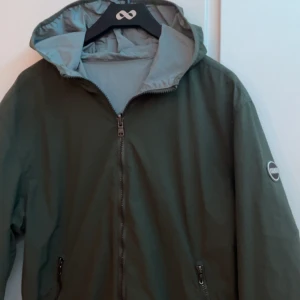 Colmar vindjacka - Säljer en stilren olivgrön vindjacka från Colmar med huva och dragkedja framtill. Jackan har två fickor med dragkedja och en diskret logotyp på ärmen. Perfekt för blåsiga dagar och enkel att matcha med olika outfits.