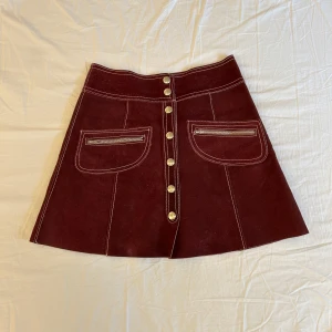 Vinröd mockakjol med dragkedjor och knappar - Säljer en vinröd vintage kjol i mocka med markerade vita sömmar. Framtill finns två fickor med dragkedjor och en rad med silvriga knappar. 