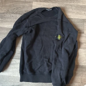 Svart sweatshirt från Stone Island - Svart sweatshirt från Stone Island med klassisk rund halsringning och ikonisk logotyp-patch på vänster ärm. Tröjan har långa ärmar och ribbade muddar vid ärmslut och nederkant. Perfekt för en avslappnad och stilren look. Den har tyvärr hål vid armarna vid baksidan som inte syns när man har på sig den men de gör att fixa genom att sy