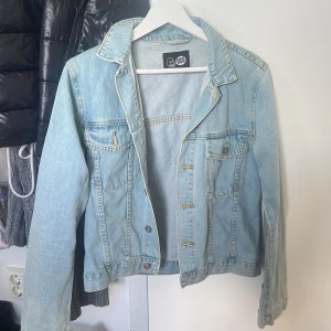Ljusblå jeansjacka - Säljer en klassisk ljusblå jeansjacka med krage och bröstfickor med knappar. Jackan har långa ärmar och stängs med knappar framtill. Perfekt att slänga över en hoodie eller t-shirt för en avslappnad look. Den är i storlek M men är väldigt liten i storleken så skulle säga mer XS/S