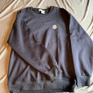 Svart sweatshirt från Moncler - Säljer en svart sweatshirt från Moncler med rund halsringning och Moncler-logga på bröstet. Tröjan har ribbade muddar vid ärmslut och nederkant. Enkel och stilren design som passar till mycket.