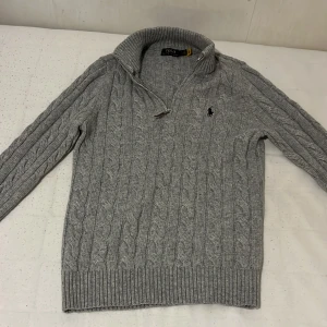 Polo Ralph Lauren halfzip  - Säljer en grå kabelstickad tröja från Polo Ralph Lauren med halv dragkedja och hög krage. Tröjan har lång ärm och klassisk broderad logga på bröstet. Perfekt för kyliga dagar och enkel att matcha med det mesta. Har aldrig använt den