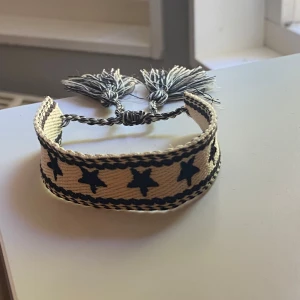 Flätat armband  - Säljer ett handgjort armband i beige tyg med blå stjärnmönster och flätade kanter. Armbandet har justerbar knytning med två stora tofsar i ändarna. Perfekt accessoar för att ge din stil en unik touch.