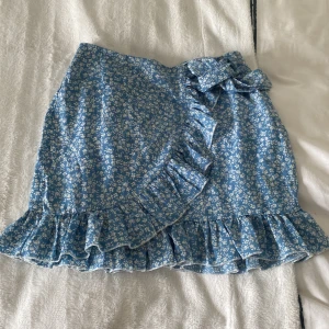 Blå blommig omlottkjol med volang från SHEIN - Supersöt blå omlottkjol från SHEIN med vitt blommönster, volangkant och en dekorativ rosett i midjan. Kjolen har dragkedja i sidan och är perfekt för vår och sommar.