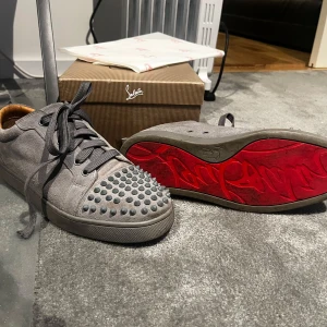 Gråa sneakers med nitar från Christian Louboutin - Säljer ett par gråa sneakers från Christian Louboutin med ikonisk röd sula och nitar framtill. Skorna har snörning och är tillverkade i mocka. Perfekta för dig som vill sticka ut med en unik detalj på fötterna.