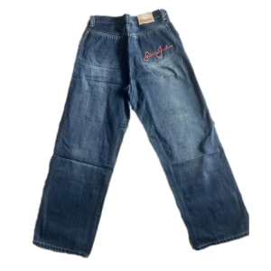 Blå jeansbyxor från Sean John med broderad logga - Säljer ett par blå jeansbyxor från Sean John med röd broderad logga på bakfickan. Byxorna har raka ben och klassisk femficksdesign. Perfekta för dig som gillar streetwear! Inte använda och i perfekt skick! 