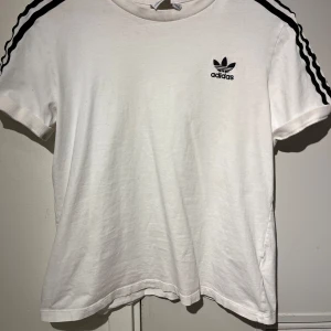 Vit t-shirt från Adidas med svarta ränder - Vit t-shirt från Adidas med klassiska svarta ränder längs axlarna och svart Adidas-logga på bröstet. Rund halsringning och korta ärmar. Perfekt för en sportig och avslappnad stil.