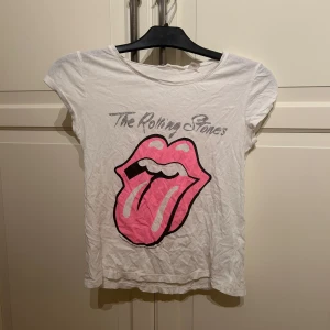 Vit Rolling Stones t-shirt med rosa tryck - Vit t-shirt med klassiskt Rolling Stones-tryck i rosa och svart framtill. Rund halsringning och korta ärmar. Perfekt för dig som gillar bandtröjor och vill sticka ut med en ikonisk design. I strl xs