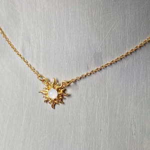 Guldhalsband med solhänge och vit sten - Snyggt halsband i guld med ett solformat hänge och en vit rund sten i mitten. Kedjan är tunn och justerbar baktill för perfekt passform. Perfekt accessoar för att lyfta din stil med en unik detalj.