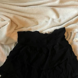 Svart volangkjol med resårmidja - Säljer en svart kortkjol med bred resår i midjan och flera lager volanger. Kjolen har en luftig och flowy känsla, perfekt att styla med en enkel topp eller hoodie. Passar dig som gillar en feminin och trendig look.