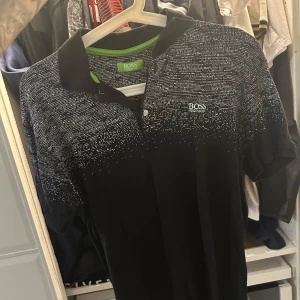 Svart Boss t-shirt med pixelmönster piké  - Svart t-shirt från Hugo Boss med vit pixelmönstrad överdel och klassisk krage. T-shirten har korta ärmar, två knappar vid halsen och Boss-logga på bröstet. Insidan av kragen har en grön detalj som ger extra edge. Perfekt för dig som gillar stilren design med twist.