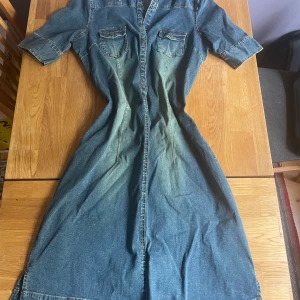 Jeansklänning från Hennes, storlek 44 - Säljer en lång jeansklänning från Hennes i storlek 44. Klänningen har korta ärmar, knappar hela vägen framtill och två bröstfickor med lock. Den är tillverkad i klassiskt blått jeanstyg med lätt tvättad effekt. Är stretchig i materialet.