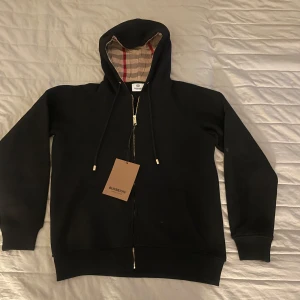 Svart Burberry hoodie  - Svart hoodie från Burberry i storlek M med klassisk rutmönstrad huva i beige och röda toner. Den är aldrig använd lappen är på 