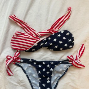 Randig och stjärnmönstrad bikini från Lebole - Cool bikini från Lebole med bandeau-topp och knyt i sidorna. Toppen har röda och vita ränder, medan både topp och trosa har marinblå botten med vita stjärnor. Materialet är mjuk polyester och elastan, perfekt för stranden. Snygg och unik design som sticker ut!