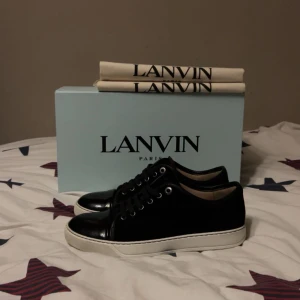 Svarta sneakers från Lanvin i mocka - Snygga svarta sneakers från Lanvin med ovandel i mocka och detaljer i blankt skinn vid tån. Skorna har vita platta sulor och svarta snören, samt metallfärgade öljetter. Perfekta för dig som gillar stilrena och lyxiga sneakers med exklusiv känsla.