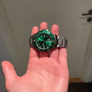 Grön klocka från Regal med stållänk - Snygg klocka från Regal med grön urtavla och silverfärgad boett. Klockan har en robust länk i metall och tydliga siffror på vridringen. Perfekt accessoar för dig som vill sticka ut med en cool färg på handleden.