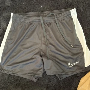 Nike Dri-Fit svarta träningsshorts - Svarta Nike Dri-Fit shorts med vita sidor och snörning i midjan. Perfekta för träning eller sport, med lätt och snabbtorkande material. Klassisk Nike-logga broderad framtill. Sitter skönt och har elastisk midja för extra komfort.