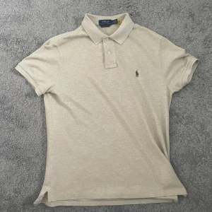 Beige pikétröja från Polo Ralph Lauren - Snygg beige pikétröja från Polo Ralph Lauren i bomull. Klassisk krage med två knappar och broderad logga på bröstet. Modellen är custom slim fit. Den är knappt använd storlek m men passar troligen S också. 