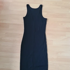 Svart ribbad långklänning - Säljer en svart långklänning med ribbad struktur och v-ringad rygg. Klänningen är ärmlös med breda axelband och har en slim passform som framhäver siluetten. Perfekt för dig som gillar minimalistisk och stilren design.